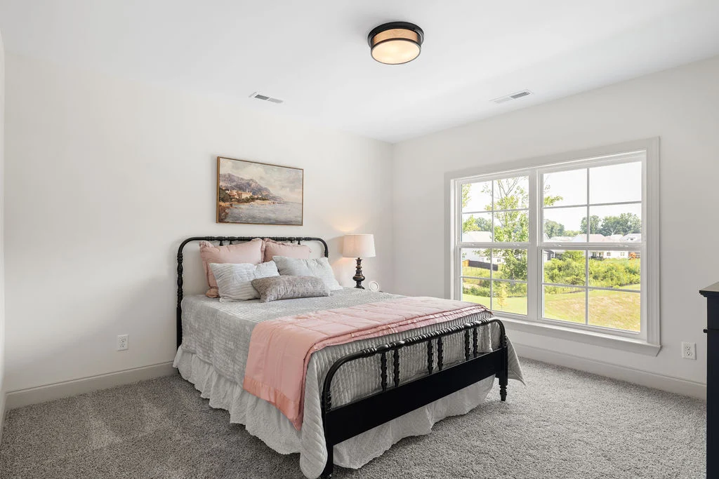 Bright bedroom with large window, pink and gray bedding, elegant lamp, and landscape artwork. Built by Pratt Home Builders in Chattanooga TN.