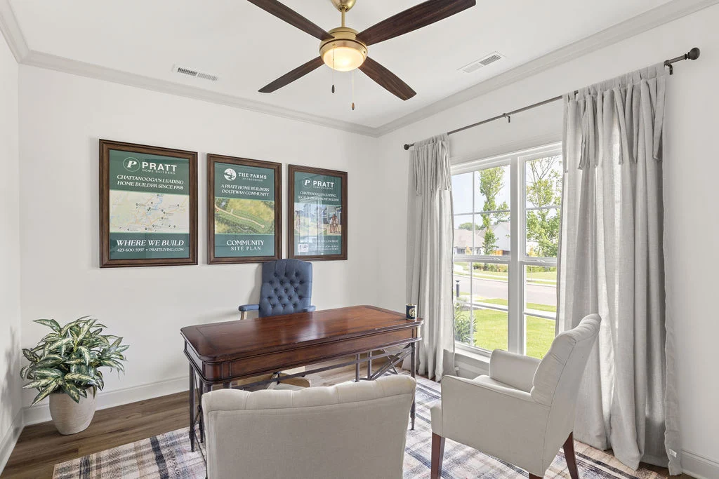 Home office with wood desk, beige chairs, window view, and Pratt Home Builders in Chattanooga TN promotional posters on the wall.