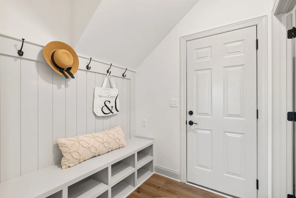 Mudroom with white bench and cubbies, hooks holding a straw hat and tote, near door. Designed by Pratt Home Builders in Chattanooga TN.