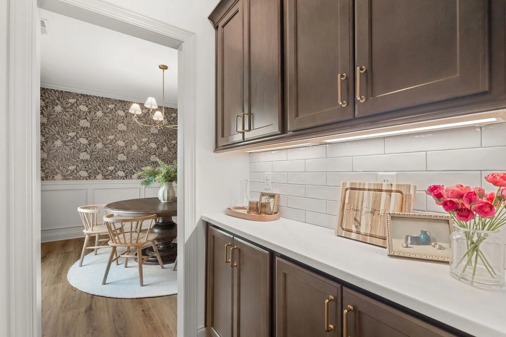 Kitchen hallway view with dark cabinets, white tiles, and red flowers; dining room has floral wallpaper and wooden chairs. Pratt Home Builders, Chatta