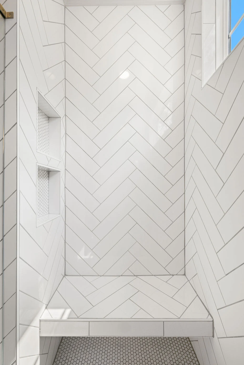 White herringbone tile shower with built-in shelves, designed by Pratt Home Builders in Chattanooga TN. Bright light from a window.