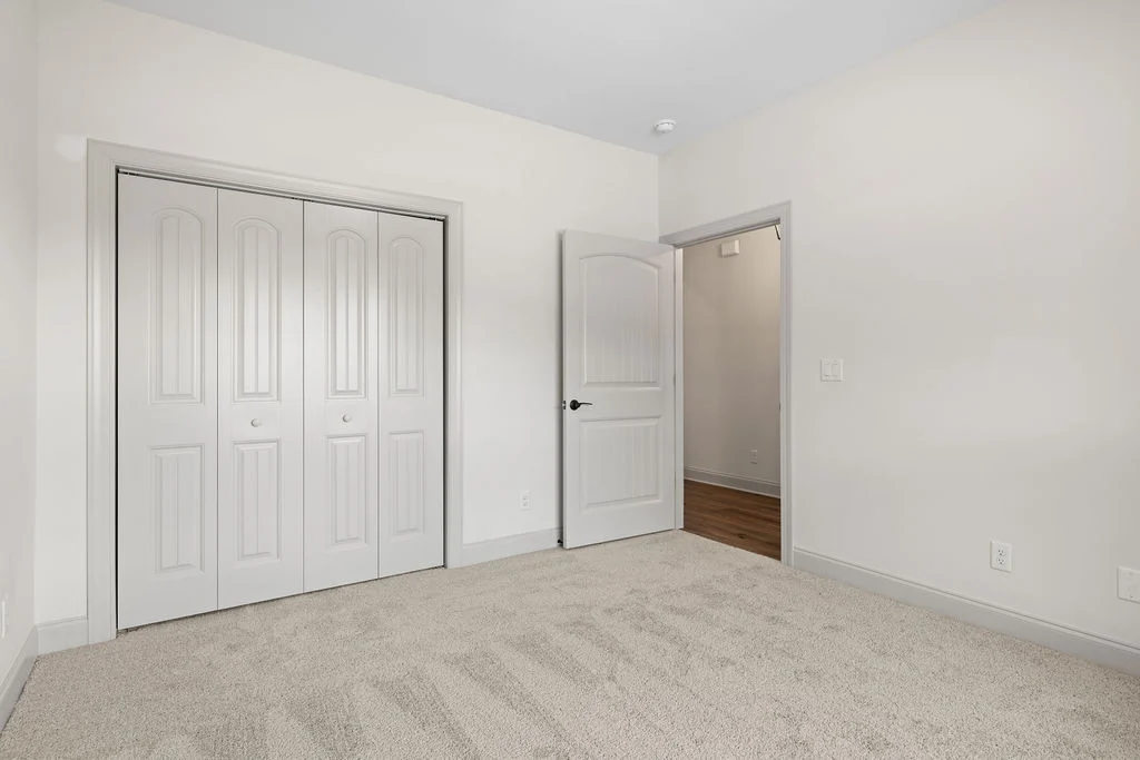 Empty room with beige carpet, closed double closet doors, and an open doorway leading to a hardwood hallway, built by Pratt Home Builders in Chattanoo