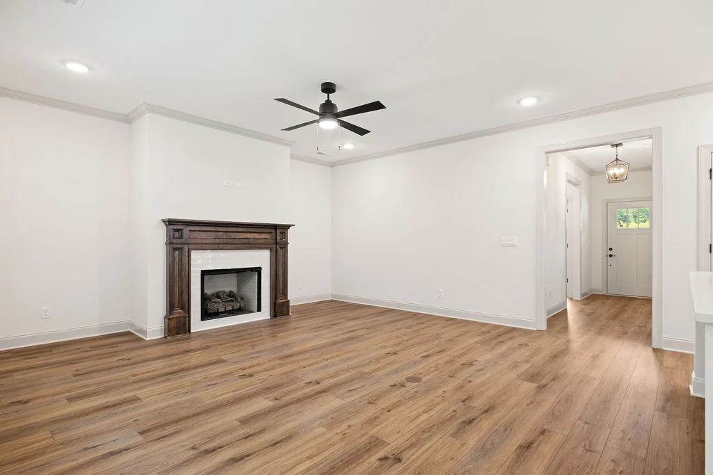 Spacious living room with wooden floors, modern ceiling fan, and elegant fireplace. Hallway leads to entrance. Built by Pratt Home Builders in Chattan