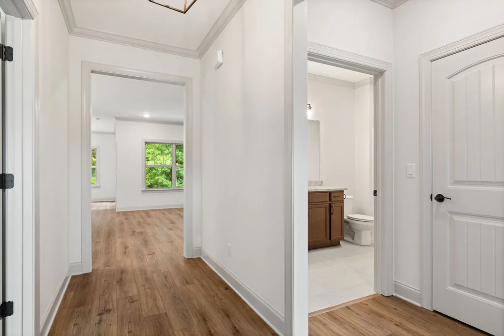 Hallway with wooden floor, leading to a bright room with large windows and a bathroom featuring brown cabinetry by Pratt Home Builders in Chattanooga