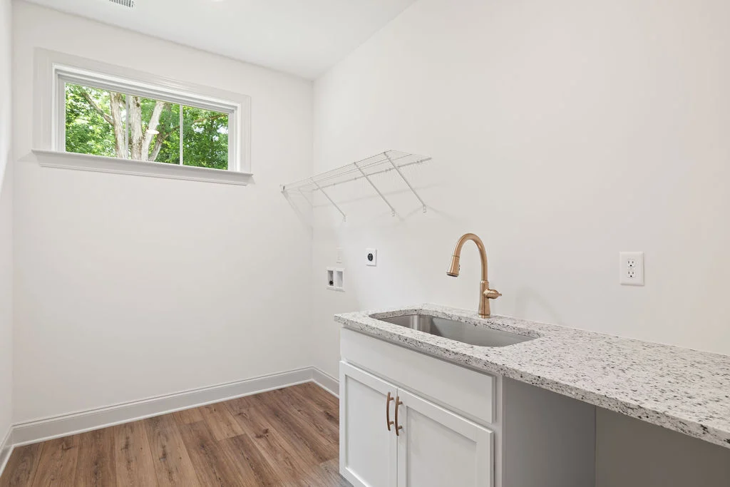 Laundry room with granite countertop, white cabinets, and gold faucet by Pratt Home Builders in Chattanooga TN, with a window view of trees.