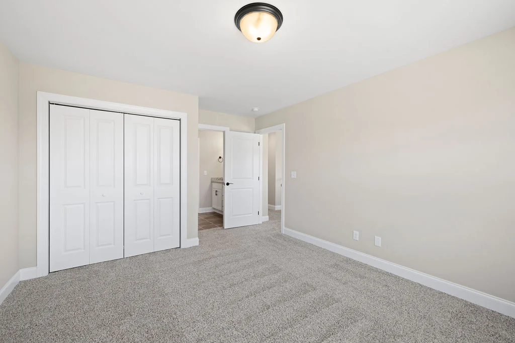 Empty bedroom with beige walls, carpet flooring, closet with white double doors, and open door to a bathroom. Built by Pratt Home Builders in Chattano