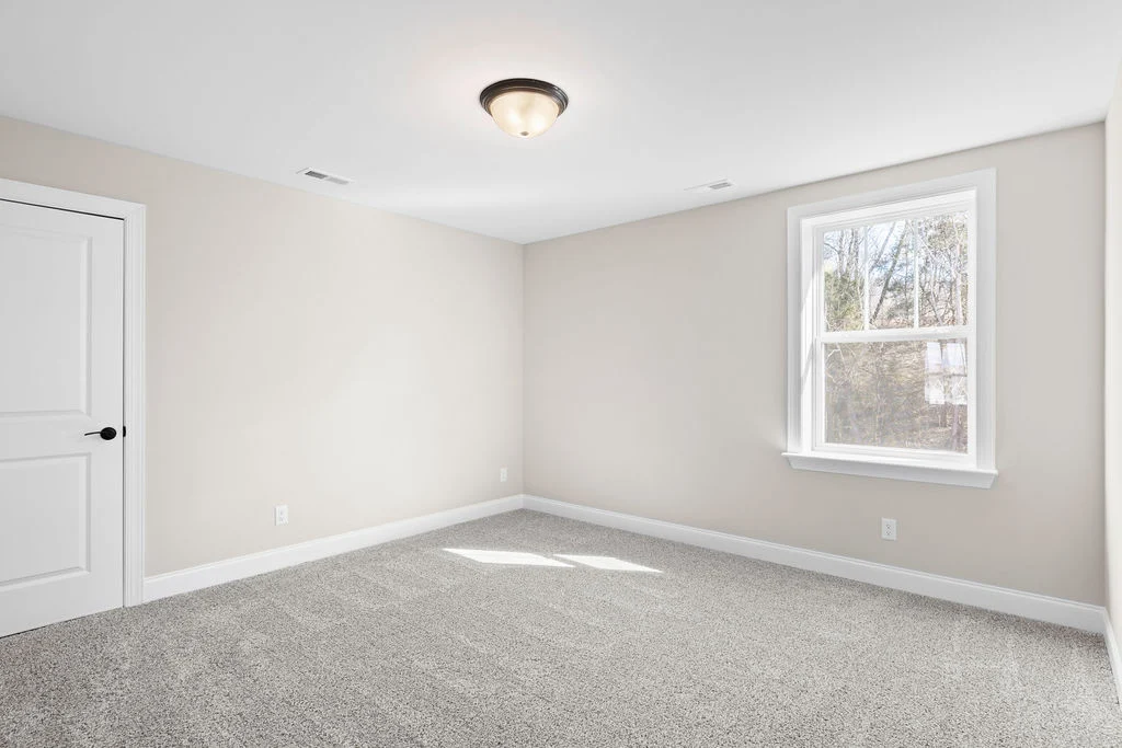 Bright, empty room with beige walls, carpeted floor, large window, and white door. Built by Pratt Home Builders in Chattanooga TN.