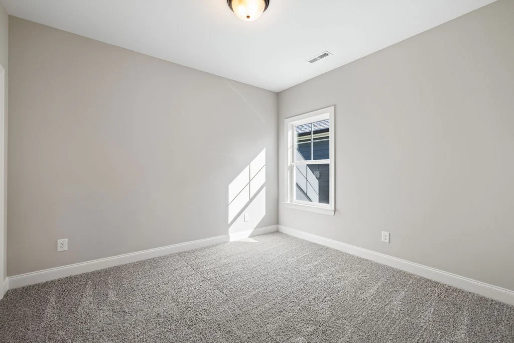 Empty room with beige walls, gray carpet, and single window. Ceiling light casts shadows on the floor. Built by Pratt Home Builders in Chattanooga TN.