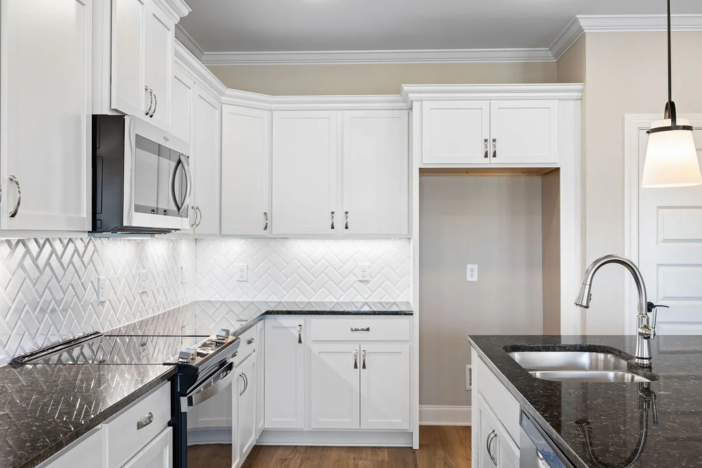 Modern kitchen with white cabinets and herringbone backsplash, featuring sleek appliances and dark granite countertops by Pratt Home Builders in Chatt