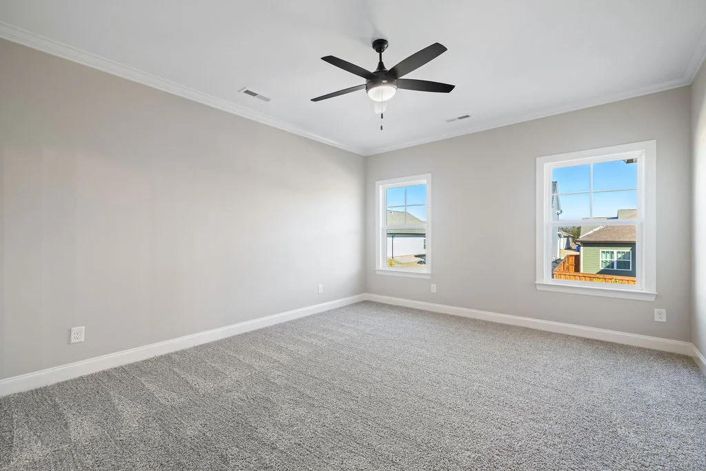 Spacious primary bedroom with gray carpet, ceiling fan, and two windows in a Pratt Home Builders house in Chattanooga, TN.