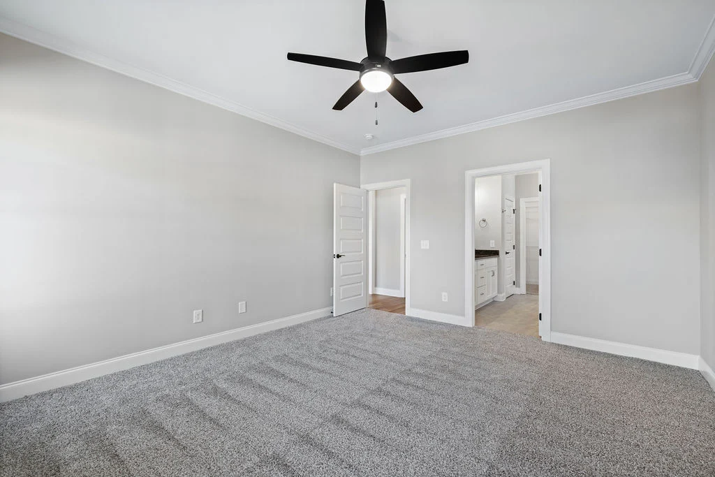 Spacious master bedroom with gray carpet, white walls, ceiling fan, and door leading to bathroom, by Pratt Home Builders in Chattanooga TN.