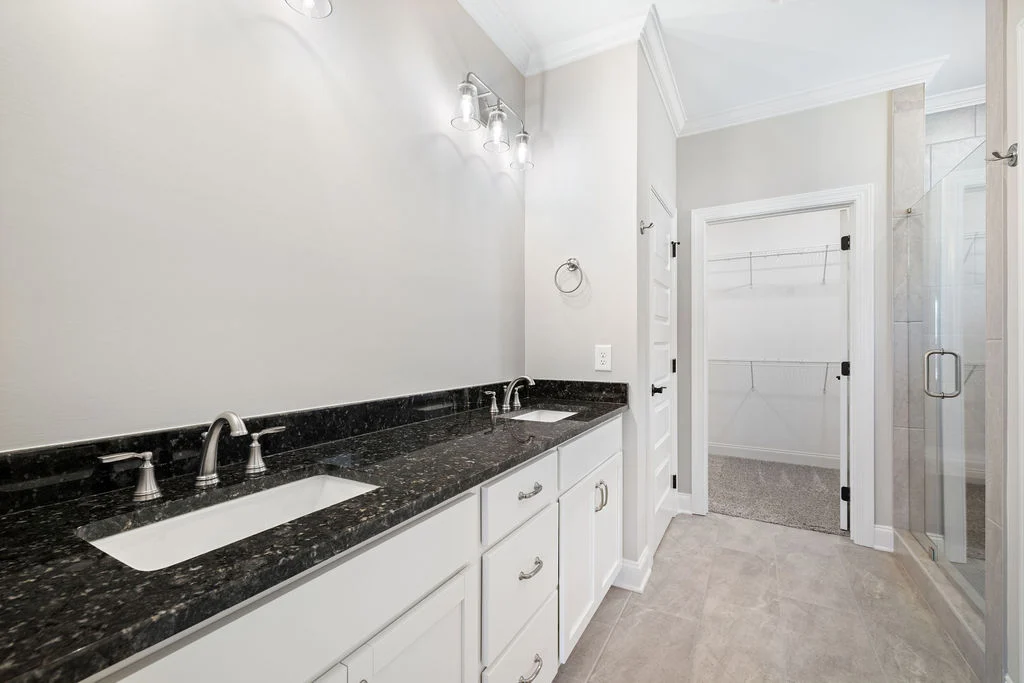 Modern primary bathroom with white cabinets and black granite countertop. Double sinks, large mirror, glass shower, walk-in closet by Pratt Home Builders, Cha