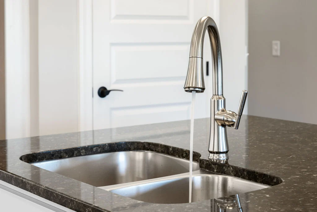 Stainless steel faucet over granite countertop in modern kitchen by Pratt Home Builders in Chattanooga TN. Water flows from the faucet.