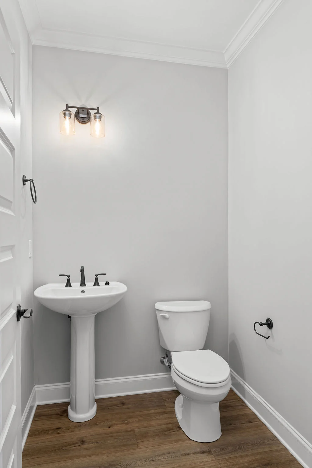 Modern bathroom with white pedestal sink, toilet, and wooden floor by Pratt Home Builders in Chattanooga TN. Black fixtures and dual bulb lighting.