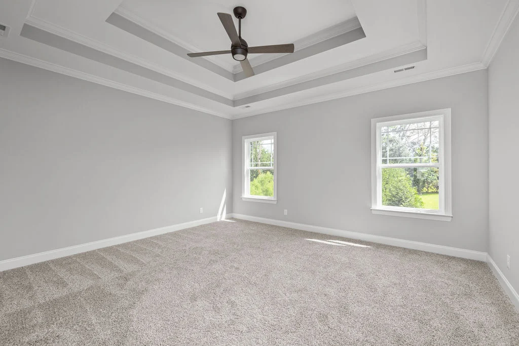 Spacious, carpeted bedroom with tray ceiling and fan, featuring two bright windows. Designed by Pratt Home Builders in Chattanooga, TN.