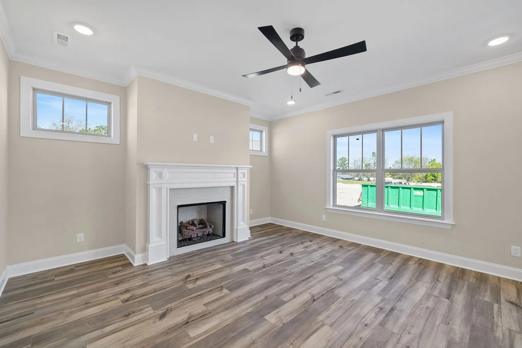 Spacious living room with hardwood floors, a ceiling fan, large windows, and a white fireplace, built by Pratt Home Builders in Chattanooga TN.