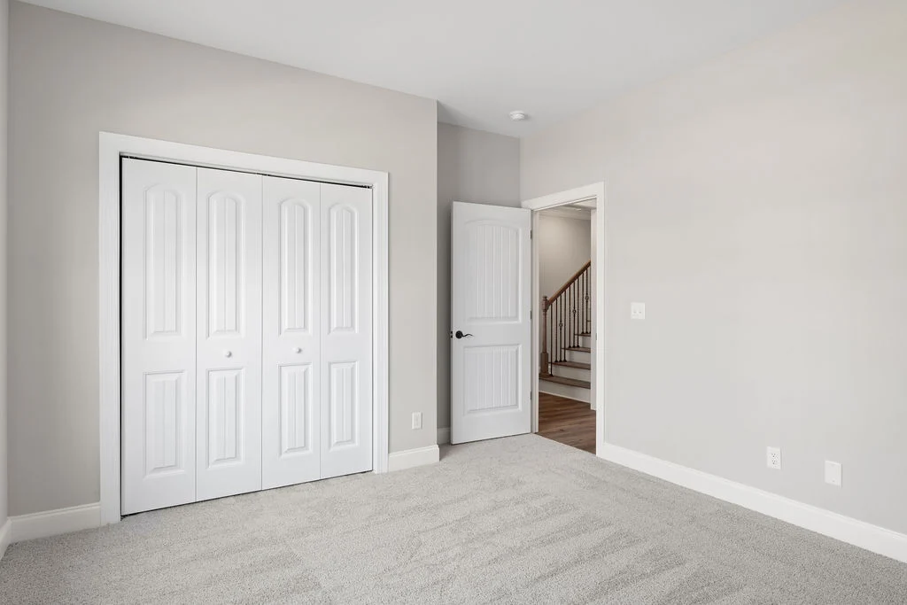 Empty room with white double closet doors, open door leading to stairs. Neutral tones, carpeted floor. Pratt Home Builders in Chattanooga TN.