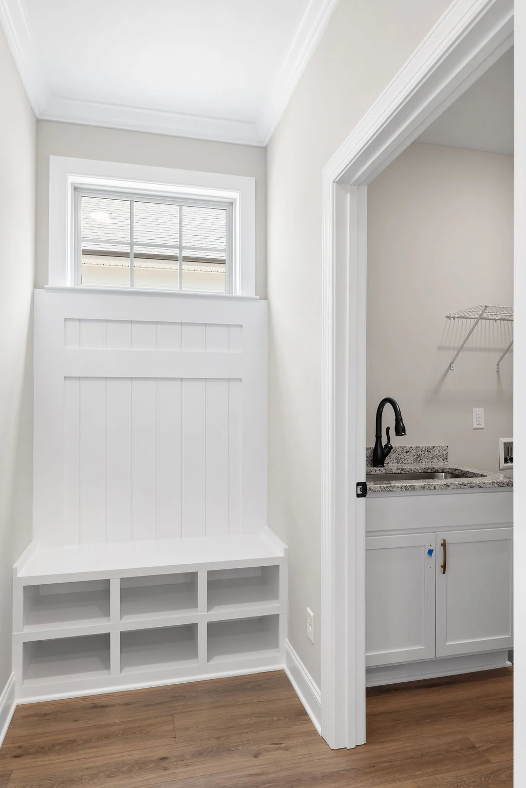 White mudroom bench with cubbies under window, adjacent to a laundry room with granite counter and black faucet. Designed by Pratt Home Builders in Ch