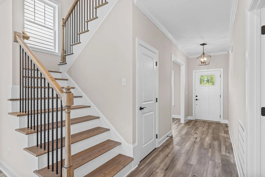Bright hallway with wooden stairs and white walls, featuring a black metal railing. Entry door with glass panel. Pratt Home Builders in Chattanooga TN