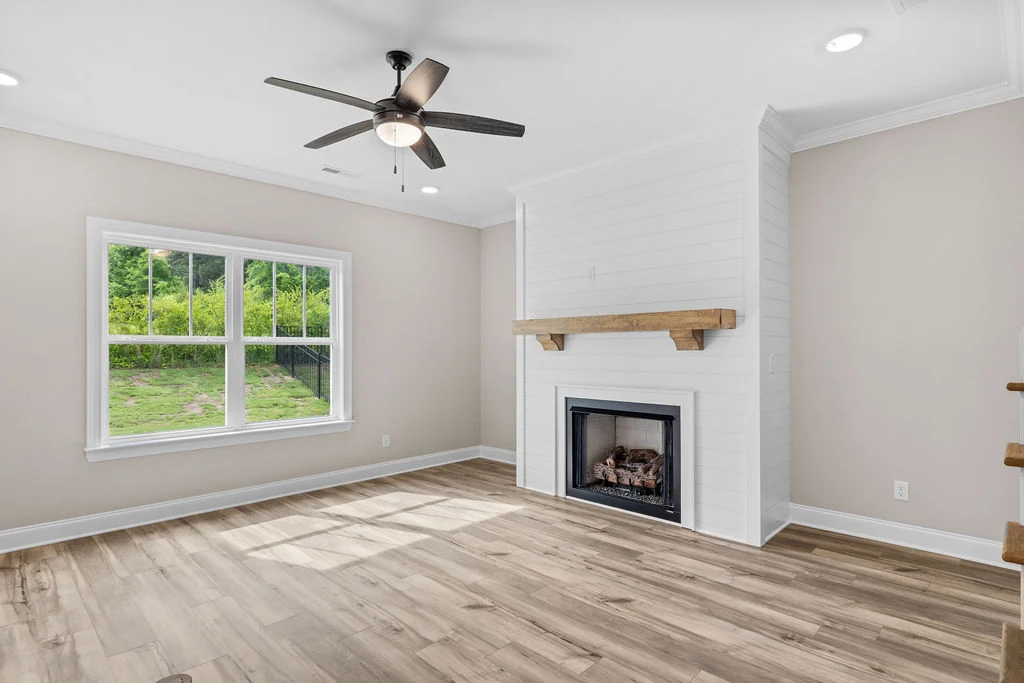Spacious living room with wooden flooring, shiplap fireplace, ceiling fan, and large window by Pratt Home Builders in Chattanooga TN.