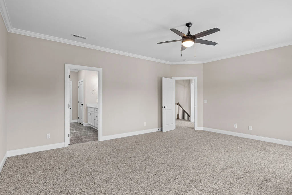 Spacious beige bedroom with plush carpet, ceiling fan, and open doors to bathroom and hallway. Designed by Pratt Home Builders in Chattanooga TN.