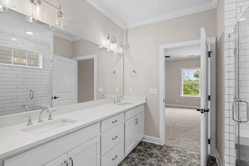 Elegant bathroom by Pratt Home Builders in Chattanooga TN, features dual sinks, glass shower, and tile flooring, with doorway leading to a carpeted ro