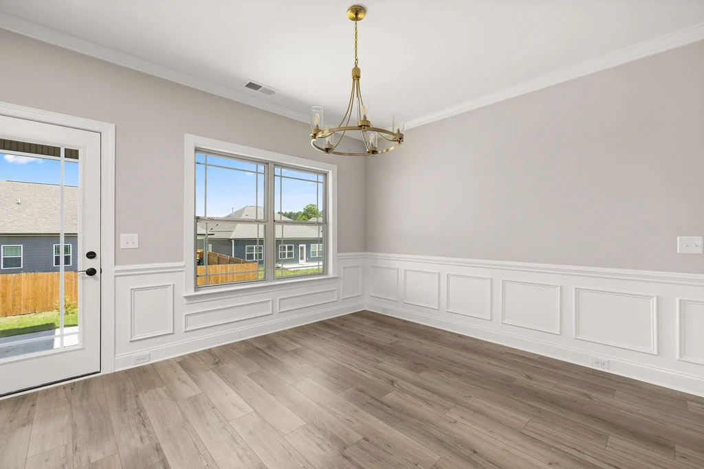 Bright dining room with wood flooring, wainscoting, and gold chandelier by Pratt Home Builders. Large window and door to backyard.