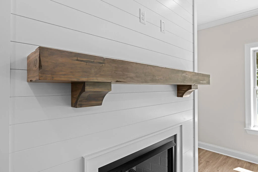 Rustic wooden mantelpiece on white shiplap wall by Pratt Home Builders in Chattanooga TN, above a black fireplace, next to a bright window.