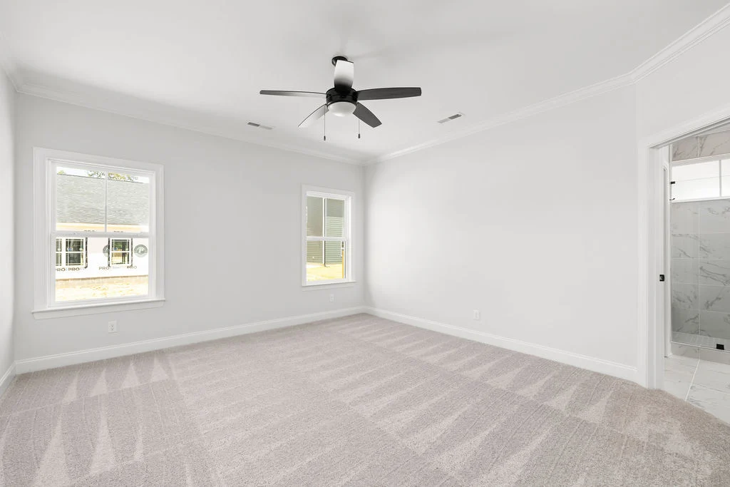 New Home Chattanooga TN. Modern bedroom with grey carpet, ceiling fan, and two windows; bathroom entrance visible. Built by Pratt Home Builders.