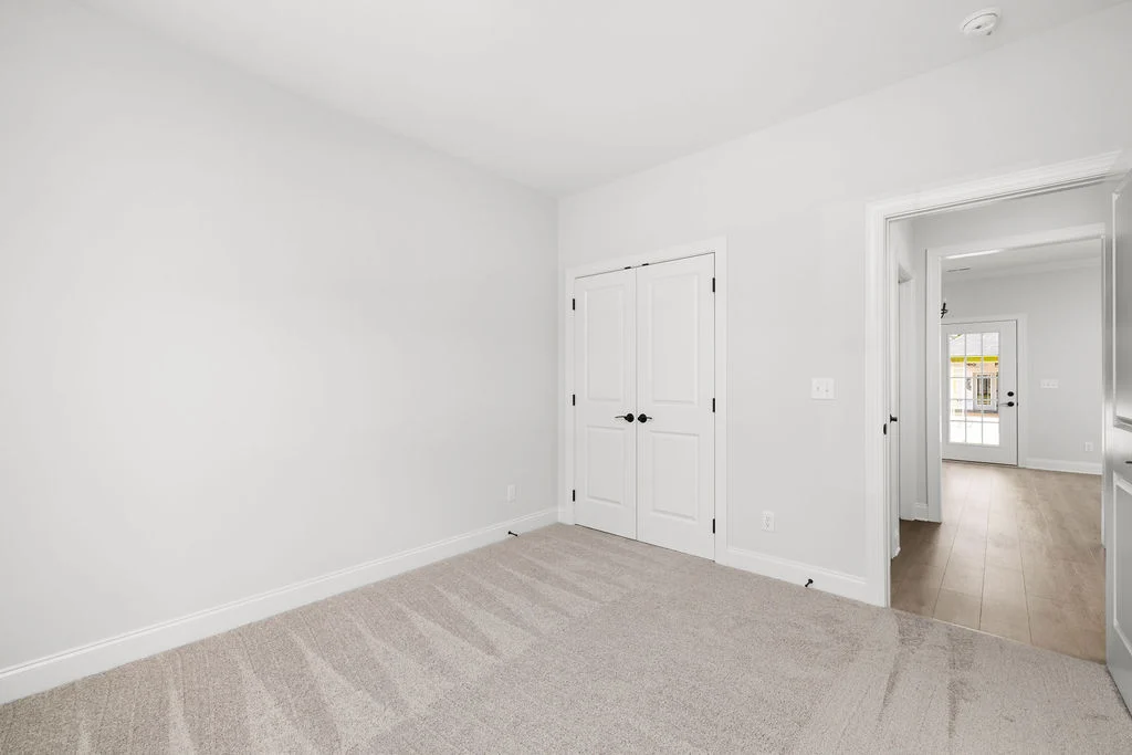 Bright, empty room with white walls, carpeted floor, and double doors. Visible hallway leading to a door with windows. Pratt Home Builders in Chattano