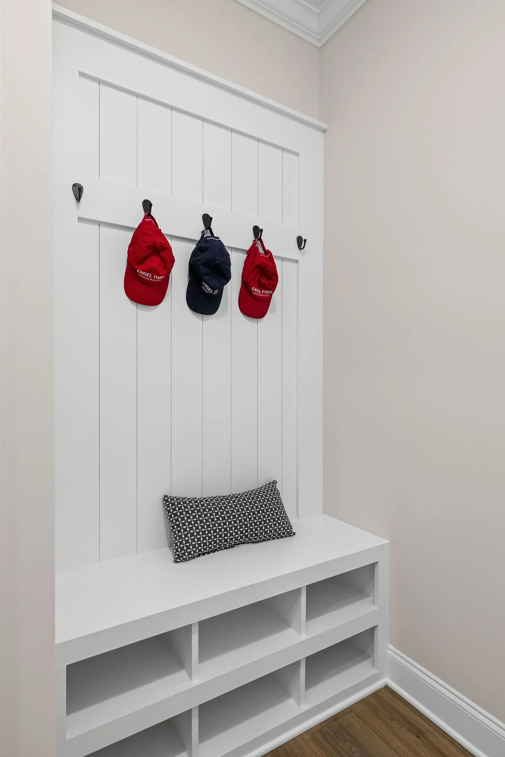 White entryway bench with hook rack, red and blue caps hanging, black and white cushion. Crafted by Pratt Home Builders in Chattanooga TN.