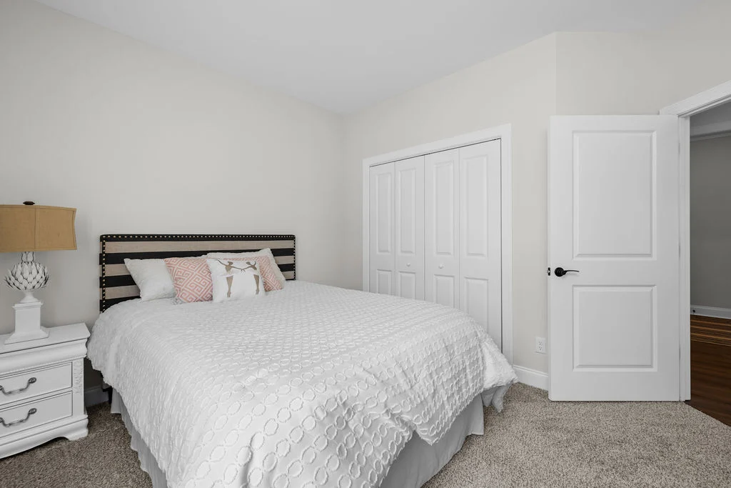 Cozy bedroom by Pratt Home Builders in Chattanooga TN, featuring white bedding, nightstand with lamp, closet, and open door.