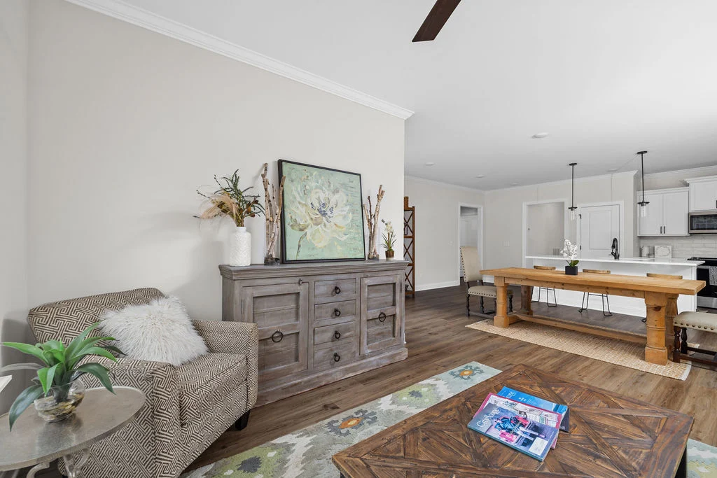 Open living room with beige walls, rustic furniture, patterned armchair, wooden dining table, and a kitchen view. Designed by Pratt Home Builders in C