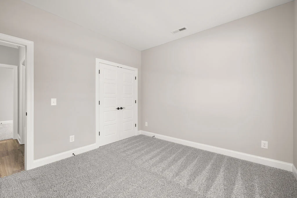 Empty room with gray carpet, beige walls, and white double doors by Pratt Home Builders in Chattanooga TN. Wooden floor visible through open doorway.