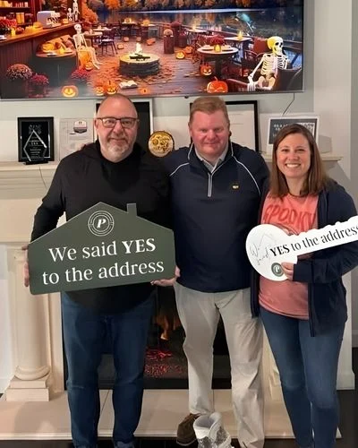 Three people smiling, holding signs that say "We said YES to the address," in a cozy room. Halloween decor present. Pratt Home Builders in Chattanooga