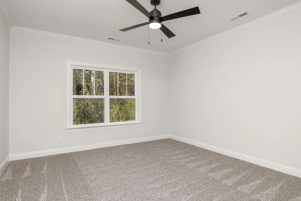 Empty room with gray carpet, white walls, ceiling fan, and large window overlooking greenery, built by Pratt Home Builders in Chattanooga TN.