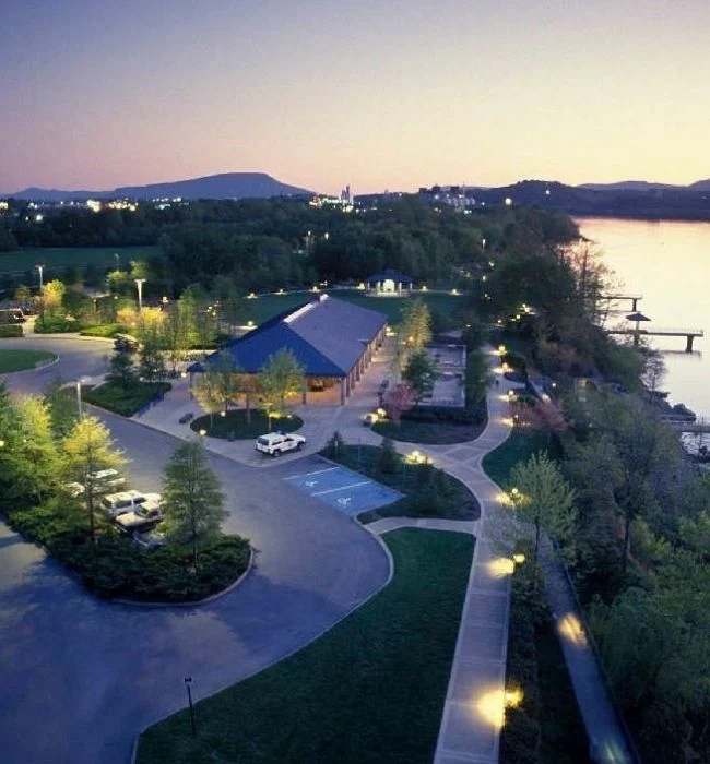 Aerial view of a serene park at dusk with a lit pathway, a building, parking area, and a river. Nearby city lights glow in the distance.