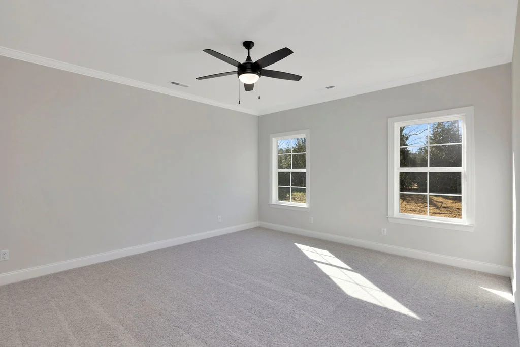 New Home Chattanooga TN. Spacious empty room with gray walls, carpet, white trim, and ceiling fan. Sunlight streams through two windows. Pratt Home Builders.
