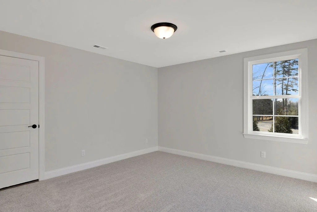 New Home Chattanooga TN. Empty, carpeted room with beige walls, white door, and window showing outdoor view; built by Pratt Home Builders.
