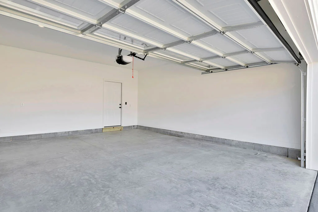 New Home Chattanooga TN. Spacious, empty two-car garage with concrete floor, by Pratt Home Builders. White walls and ceiling with open roller door.
