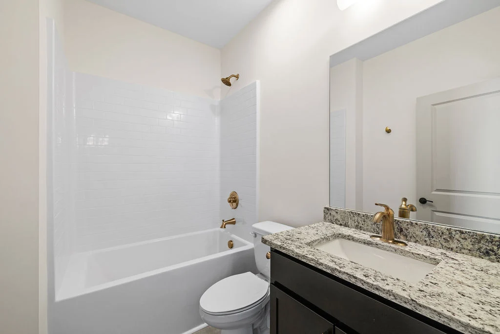 Modern bathroom by Pratt Home Builders in Chattanooga TN features granite vanity, white tub-shower combo, gold fixtures, and a large mirror.
