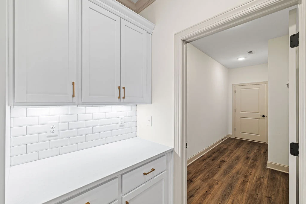 New Home Chattanooga TN. White cabinetry with gold handles next to a bright hallway by Pratt Home Builders. Wood flooring and subway tile backsplash.