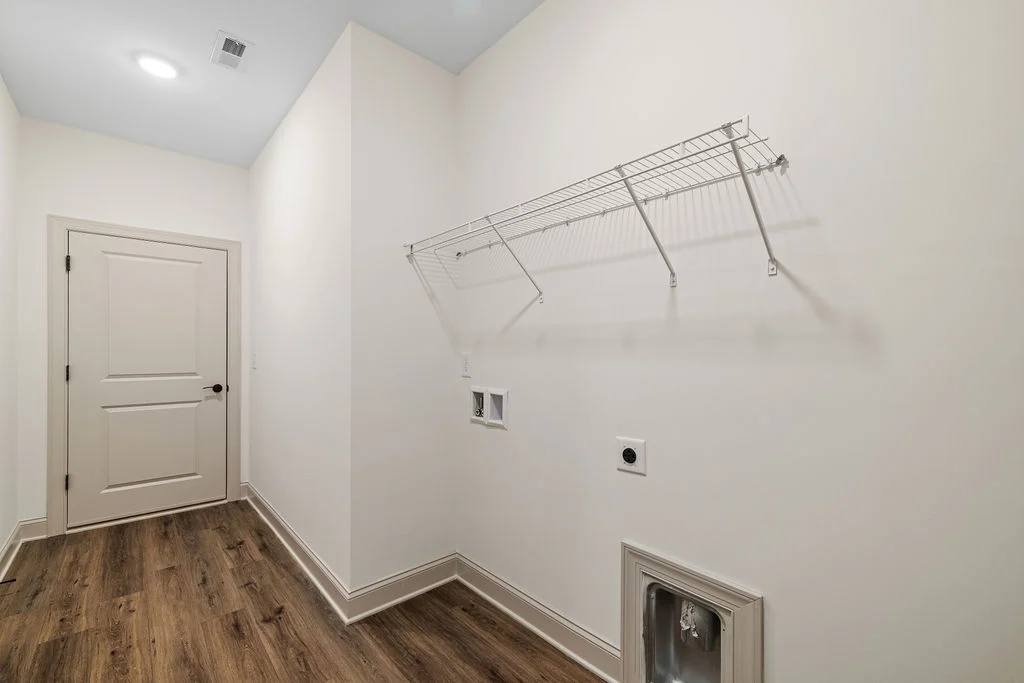 New Home Chattanooga TN. Laundry room with wire shelf, hookups, and wooden floors by Pratt Home Builders. White walls and closed door on the left.