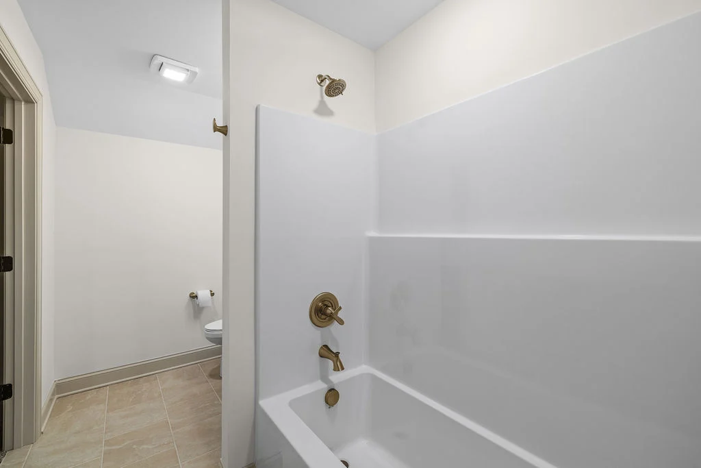 Bathroom with white tub, gold fixtures, beige tile floor, and partially visible toilet; designed by Pratt Home Builders in Chattanooga TN.