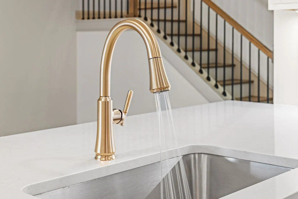 Elegant gold kitchen faucet with running water in a modern kitchen by Pratt Home Builders in Chattanooga, TN, featuring a stylish staircase backdrop.