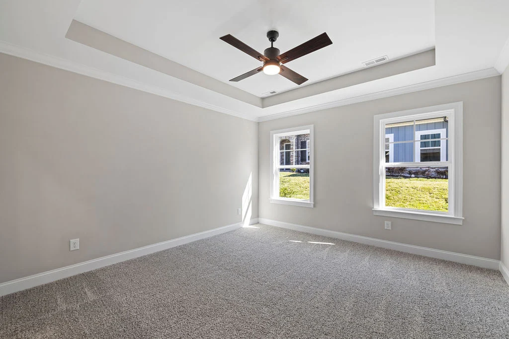 Spacious empty bedroom with gray walls, carpet, tray ceiling, and ceiling fan by Pratt Home Builders in Chattanooga TN. Bright sunlight through window