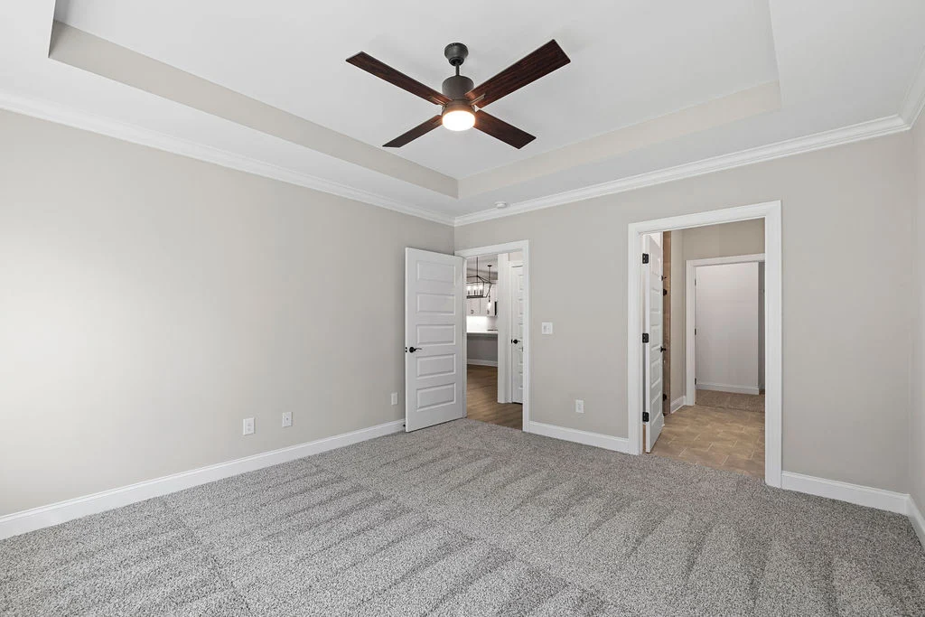 New Home Chattanooga TN. Empty room with gray carpet, tray ceiling, and ceiling fan. Two doorways lead to a kitchen and hallway. Built by Pratt Home Builders.