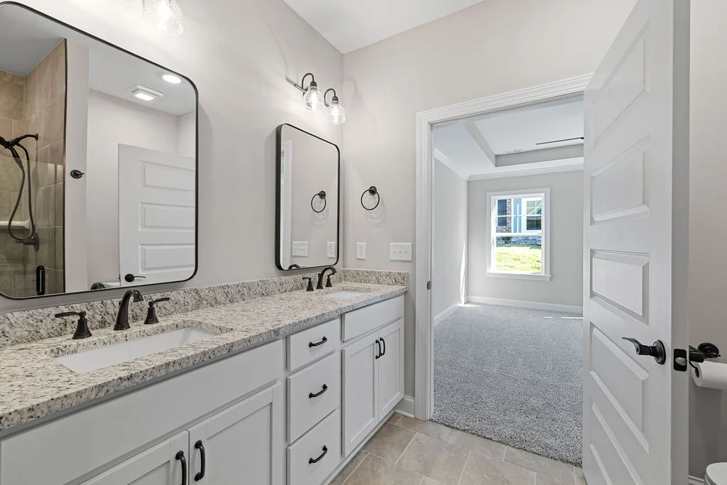 Modern bathroom with double vanity, granite counters, and black fixtures, leading to a sunlit carpeted room. Pratt Home Builders in Chattanooga TN.