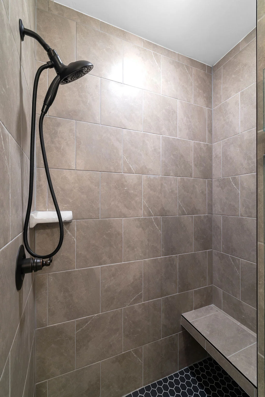 Modern shower with beige tiles, black showerhead, bench, and hexagonal floor by Pratt Home Builders in Chattanooga TN.