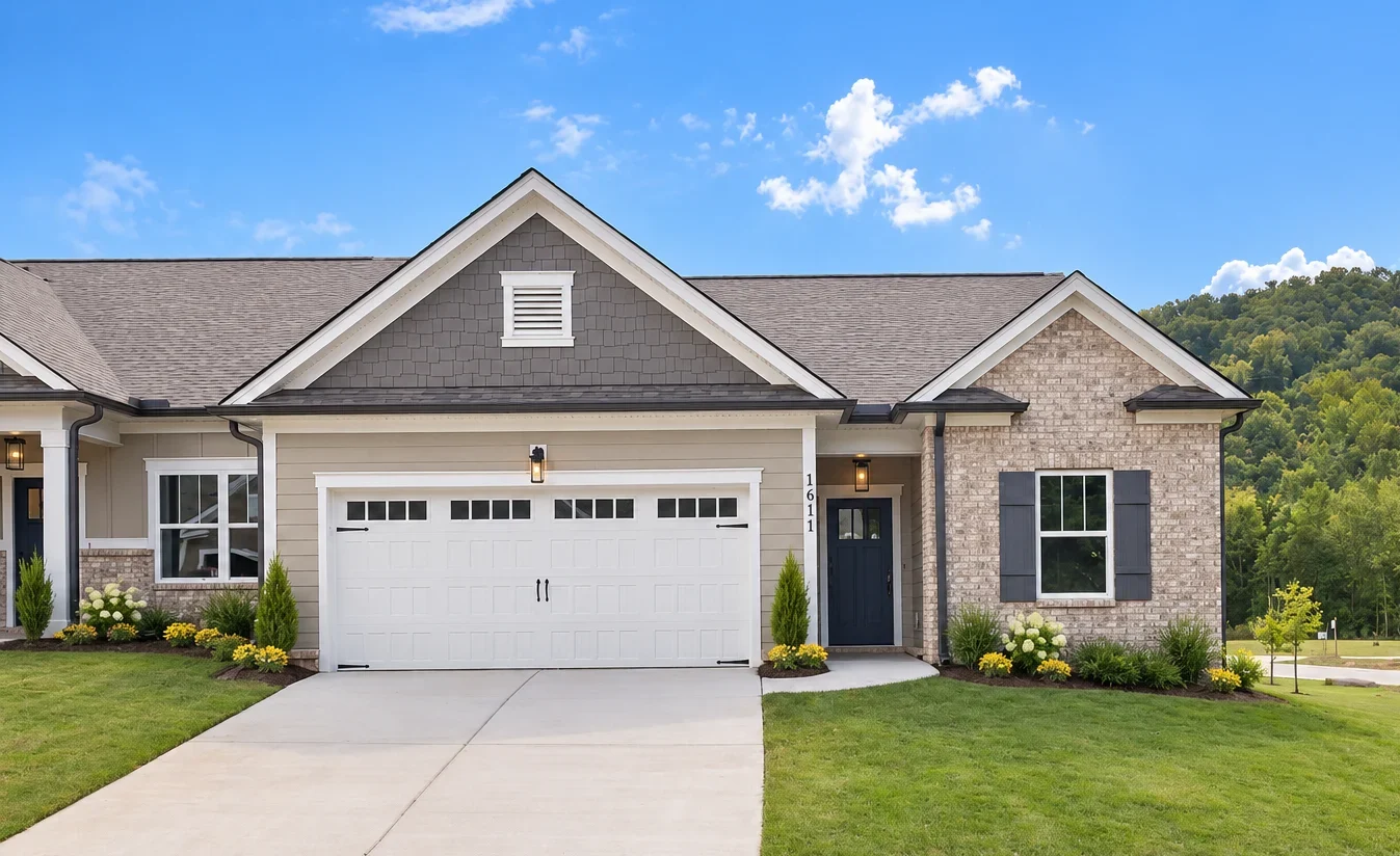 Modern single-story home with a two-car garage, gray siding, and brick accents by Pratt Home Builders in Chattanooga, TN, surrounded by lush greenery.